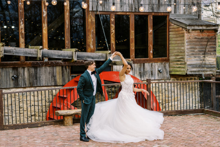 Groom spins his bride while dancing outside in front of a red waterwheel