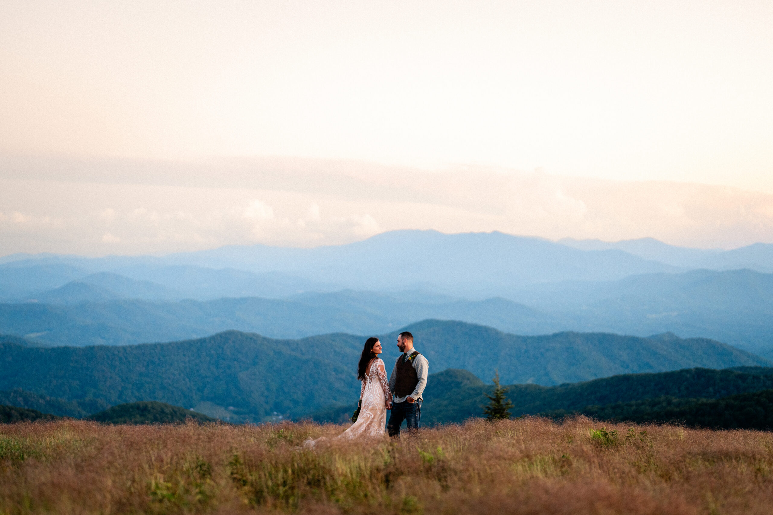 Bride and groom hold hands and stare into each others eyes while standing in a field with mountains in the background